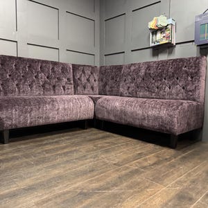 May include: A large, L-shaped, tufted sofa with a dark purple fabric upholstery. The sofa is in a room with wood floors and grey walls.