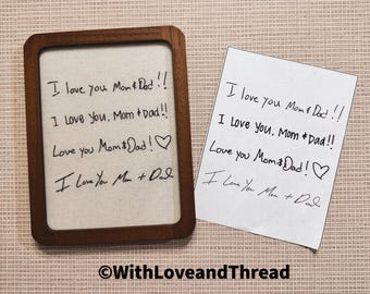 May include: A rectangular wooden frame containing a light-colored fabric insert with the black embroidered text: I love you Mom & Dad!!, I Love You, Mom & Dad!!, Love you Mom & Dad! with a heart, and I Love You Mom + Dad. Beside it is a white paper.