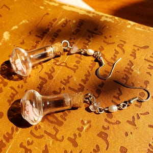 May include: A pair of unique earrings featuring small glass bottles with cork stoppers. Each bottle contains pink floral elements. The earrings have silver-colored hooks and are adorned with small beads and pearls.