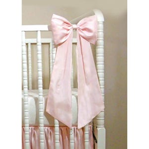May include: A white crib with a large, light pink satin bow attached to the headboard. The bow has long, flowing tails. A matching pink crib skirt with a ruffled design is visible.