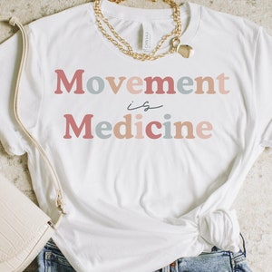 Może przedstawiać: Biała koszulka z napisem "Movement is Medicine" w r&oacute;żowych, niebieskich i beżowych literach. Koszulka jest stylizowana z dżinsowymi spodenkami i kremową torebką.