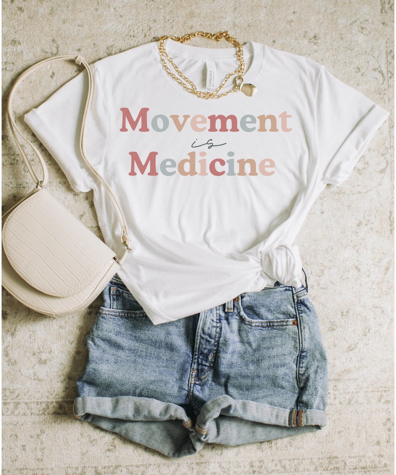 Może przedstawiać: Biała koszulka z napisem "Movement is Medicine" w r&oacute;żowych, niebieskich i beżowych literach. Koszulka jest stylizowana z dżinsowymi spodenkami i kremową torebką.