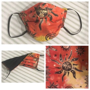 May include: A face mask with a sun design. The mask has a red, orange, and yellow background with a black and gold sun graphic. It features black elastic ear loops and a black interior lining. The mask is pleated for a comfortable fit.