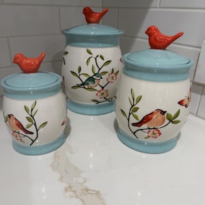 May include: A set of three ceramic canisters with light blue lids and bases. Each lid features a small orange bird figurine. The white bodies are decorated with painted branches, green leaves, pink flowers, and various birds and butterflies.