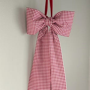 May include: A decorative bow made of red and white gingham fabric, hanging from a black rod with a red velvet ribbon. The bow is large and has a long, rectangular tail.