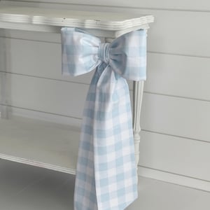 May include: A large, light blue and white gingham bow is attached to a white table. The bow is made of fabric and has a full, decorative shape. The bow's fabric hangs down from the table.