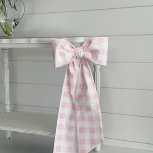 May include: A large, decorative bow made of pink and white gingham fabric. The bow is tied and draped over a white table, with the fabric cascading down. The bow adds a touch of charm and elegance to the setting.