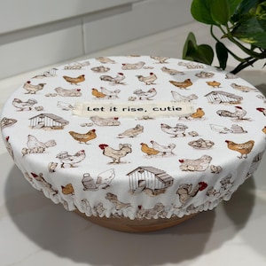 May include: A white bowl cover with a repeating pattern of chickens, chicken coops, and eggs. The cover has a ruffled edge and a fabric tag that reads "Let it rise, cutie."