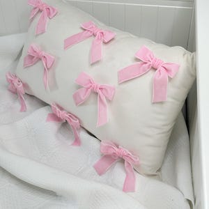 May include: A white pillow with pink velvet bows sewn on the front. The pillow is on a white bedspread.