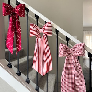 May include: Three decorative bows in red and white hang from a staircase railing. The bows feature different patterns: polka dots, stripes, and gingham. The bows are attached to a white railing with black spindles, adding a festive touch to the home decor.