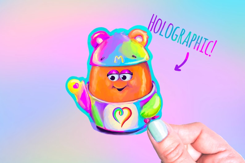 May include: A holographic sticker of a cartoon character with a rainbow colored hat and outfit. The character has a heart on its chest and is holding a heart in its hand. The sticker says "HOLOGRAPHIC!" on the top right corner.