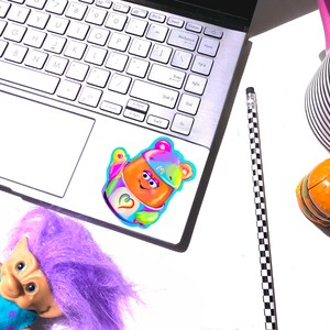 May include: A colorful sticker of a cartoon character with a rainbow hat and a heart on its chest. The sticker is on a silver laptop computer.