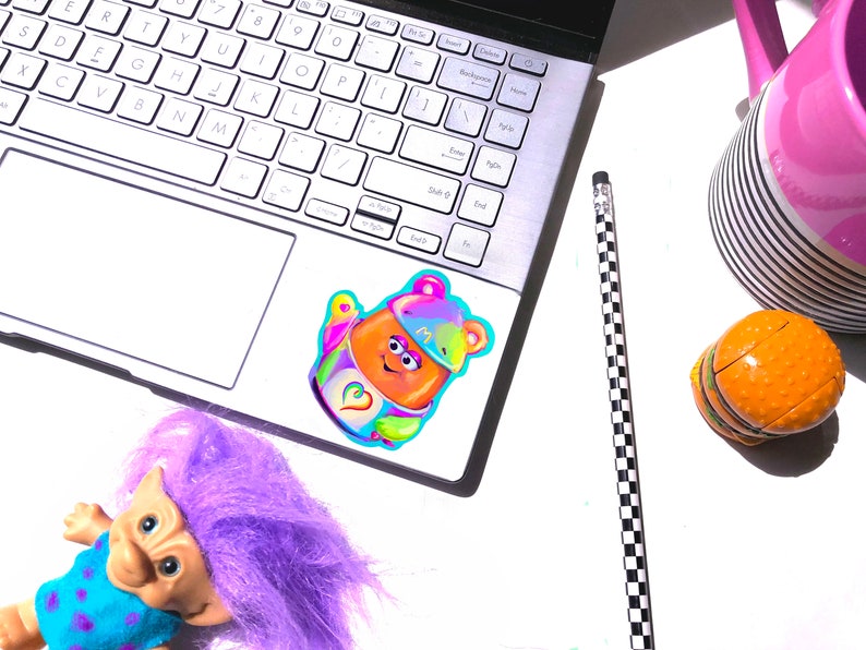 May include: A colorful sticker of a cartoon character with a rainbow hat and a heart on its chest. The sticker is on a silver laptop computer.