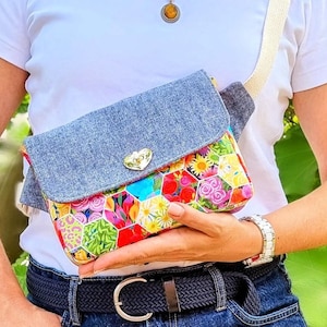 Op de afbeelding: Een schoudertas met een klep in denimtextuur en een goudkleurige hartvormige sluiting. Het hoofdgedeelte toont een levendig, veelkleurig zeshoekig patroon gevuld met diverse bloemen-, blad- en dierenprints. Het heeft een beige schouderband.