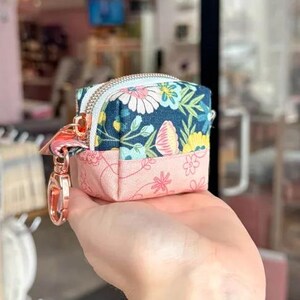 Puede incluir: Peque&ntilde;o estuche cuadrado con estampado floral y cierre de cremallera. La mitad superior del estuche es azul marino con margaritas blancas y flores amarillas. La mitad inferior es rosa con peque&ntilde;as flores rosas. Cierre dorado rosa.