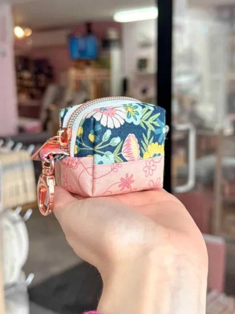 Puede incluir: Peque&ntilde;o estuche cuadrado con estampado floral y cierre de cremallera. La mitad superior del estuche es azul marino con margaritas blancas y flores amarillas. La mitad inferior es rosa con peque&ntilde;as flores rosas. Cierre dorado rosa.