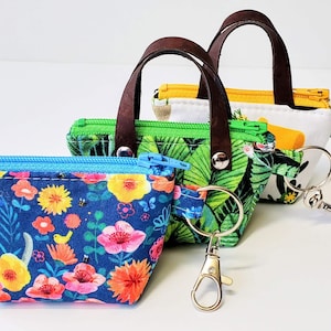 K&ouml;nnte beinhalten: Drei kleine, farbenfrohe Stoffb&ouml;rsen mit braunen Ledergriffen und Schl&uuml;sselringen. Die B&ouml;rsen zeigen Blumen-, Tropenblatt- und Katzenmuster. Jede B&ouml;rse hat einen Rei&szlig;verschluss und einen Metallverschluss zum Befestigen von Schl&uuml;sseln.