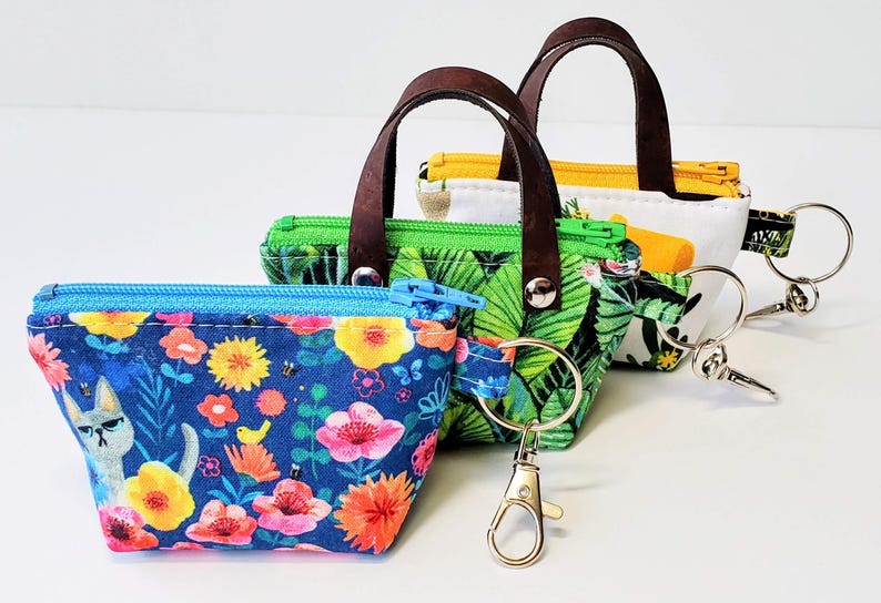 K&ouml;nnte beinhalten: Drei kleine, farbenfrohe Stoffb&ouml;rsen mit braunen Ledergriffen und Schl&uuml;sselringen. Die B&ouml;rsen zeigen Blumen-, Tropenblatt- und Katzenmuster. Jede B&ouml;rse hat einen Rei&szlig;verschluss und einen Metallverschluss zum Befestigen von Schl&uuml;sseln.