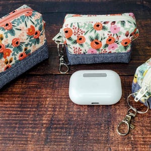 Puede incluir: Tres estuches con cremallera con estampado floral, bases de mezclilla y llaveros. Los estuches presentan un fondo crema con rosas naranjas y rosas y hojas verdes. Entre los estuches hay una funda blanca rectangular.