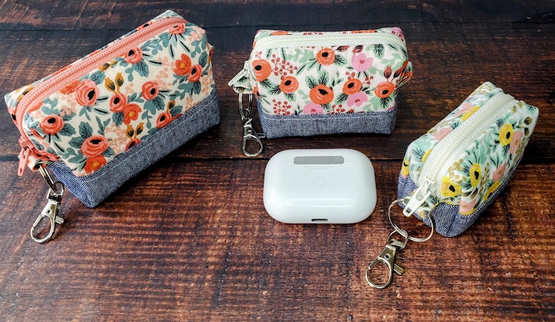 Puede incluir: Tres estuches con cremallera con estampado floral, bases de mezclilla y llaveros. Los estuches presentan un fondo crema con rosas naranjas y rosas y hojas verdes. Entre los estuches hay una funda blanca rectangular.