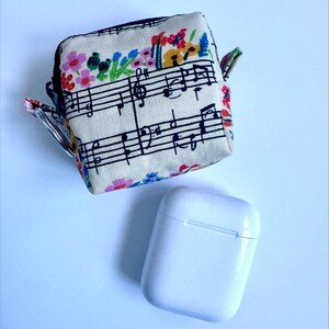 Puede incluir: Una funda cuadrada de tela beige con cremallera, con un dise&ntilde;o de pentagrama y acentos florales coloridos. Debajo hay una funda de carga de auriculares inal&aacute;mbricos blanca y rectangular.