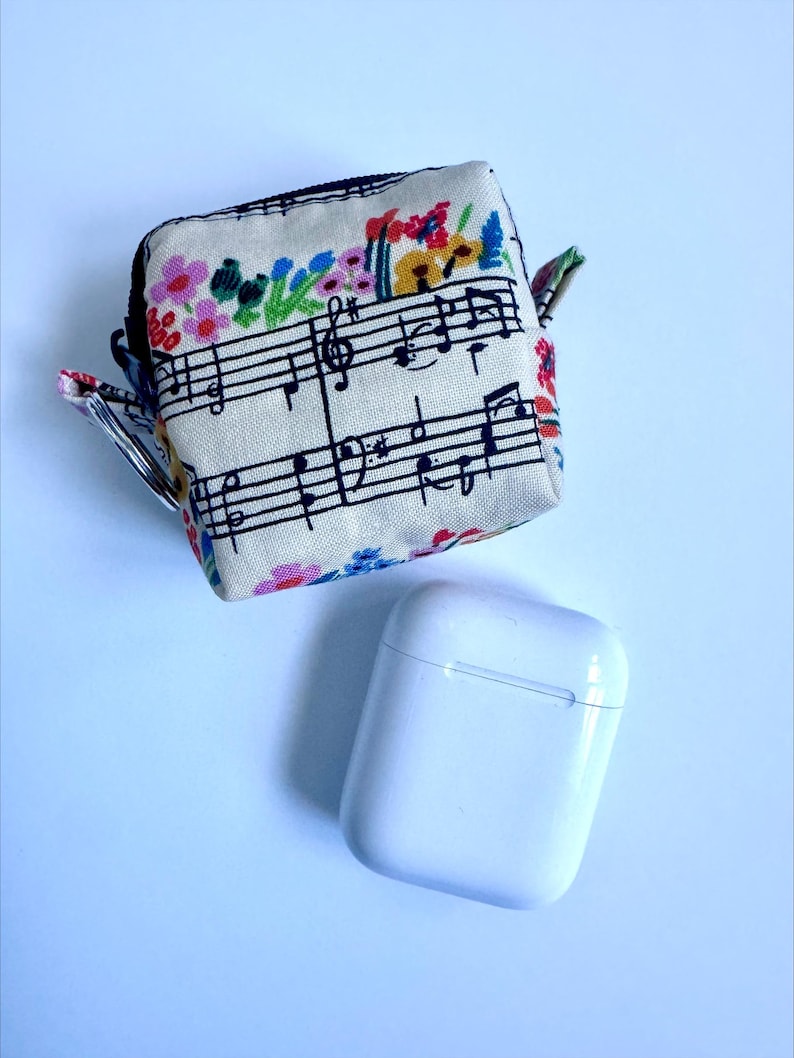 Puede incluir: Una funda cuadrada de tela beige con cremallera, con un dise&ntilde;o de pentagrama y acentos florales coloridos. Debajo hay una funda de carga de auriculares inal&aacute;mbricos blanca y rectangular.