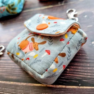 May include: A small, light blue fabric pouch with a flap closure secured by an orange snap button. The fabric features a repeating pattern of brown dogs wearing pink sweaters, red mushrooms, and gray sprigs. A silver metal lobster clasp is attached.