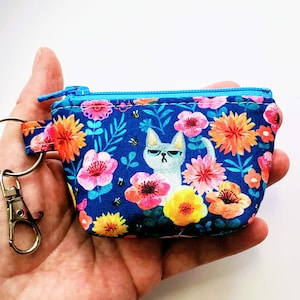 K&ouml;nnte beinhalten: Ein kleines, blaues M&uuml;nzfach mit einem Blumenmuster mit rosa, orangefarbenen und gelben Blumen und einer Illustration einer grummeligen Katze. Das Fach hat einen blauen Rei&szlig;verschluss und einen Metall-Schl&uuml;sselanh&auml;nger.