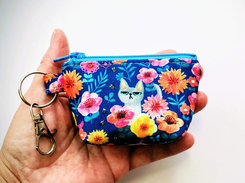 K&ouml;nnte beinhalten: Ein kleines, blaues M&uuml;nzfach mit einem Blumenmuster mit rosa, orangefarbenen und gelben Blumen und einer Illustration einer grummeligen Katze. Das Fach hat einen blauen Rei&szlig;verschluss und einen Metall-Schl&uuml;sselanh&auml;nger.