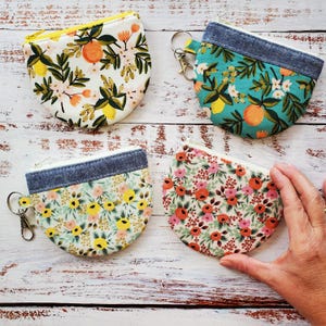 May include: Four small, half-moon shaped coin purses with floral patterns. The purses have a denim-like fabric at the top and a zipper closure. One purse is held by a hand. The patterns include oranges, lemons, and various flowers.