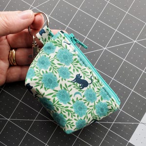 K&ouml;nnte beinhalten: Ein kleiner Geldbeutel mit floralem Muster, t&uuml;rkisblauem Rei&szlig;verschluss und Schl&uuml;sselring. Der Stoff zeigt t&uuml;rkisfarbene Blumen und gr&uuml;ne Bl&auml;tter auf wei&szlig;em Grund, mit einer marineblauen Katzen-Silhouette. Der Geldbeutel wird von einer Hand gehalten.