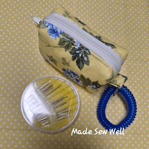 Puede incluir: Un kit de costura con estampado floral amarillo con una bolsa con cremallera blanca, un recipiente transparente de agujas y un llavero en espiral azul. El texto "Made Sew Well" es visible.