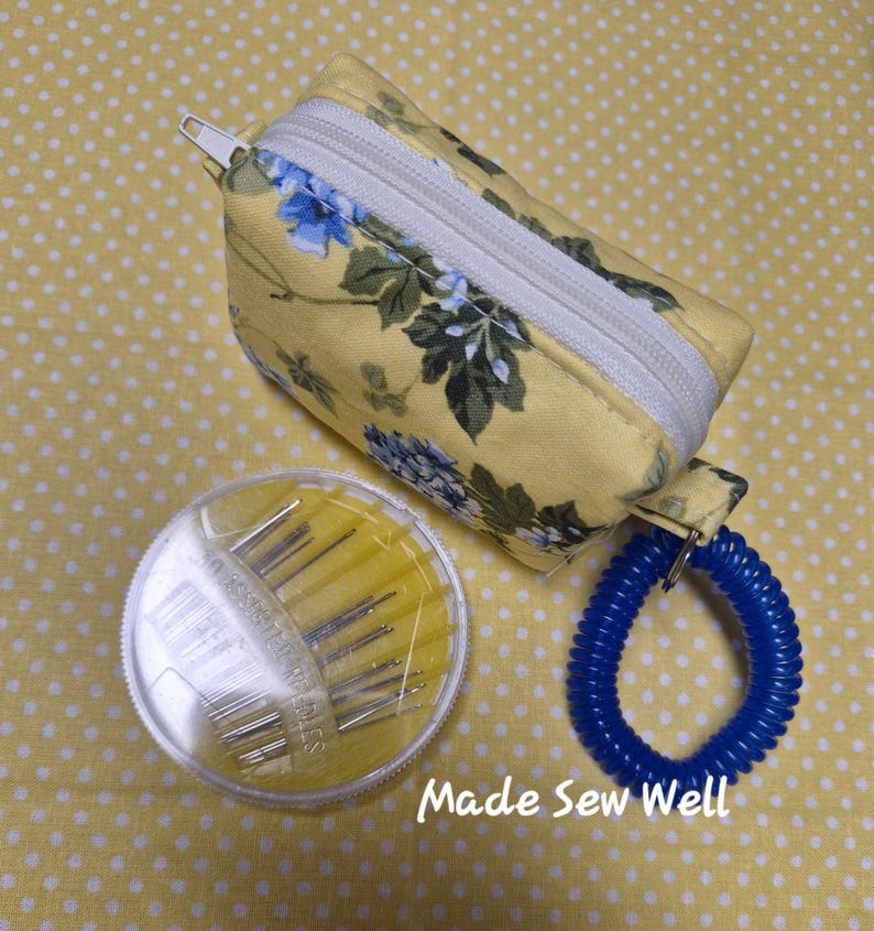 Puede incluir: Un kit de costura con estampado floral amarillo con una bolsa con cremallera blanca, un recipiente transparente de agujas y un llavero en espiral azul. El texto "Made Sew Well" es visible.