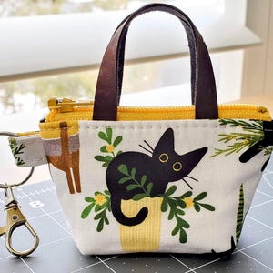 K&ouml;nnte beinhalten: Kleine Stoff-Tragetasche mit schwarzem Katzenmuster, gelbem Rei&szlig;verschluss und braunen Griffen. Die Tasche hat einen goldfarbenen Schl&uuml;sselanh&auml;nger und einen Stoffriemen mit Muster. Die Katze sitzt in einem gelben Topf mit gr&uuml;nen Bl&auml;ttern.