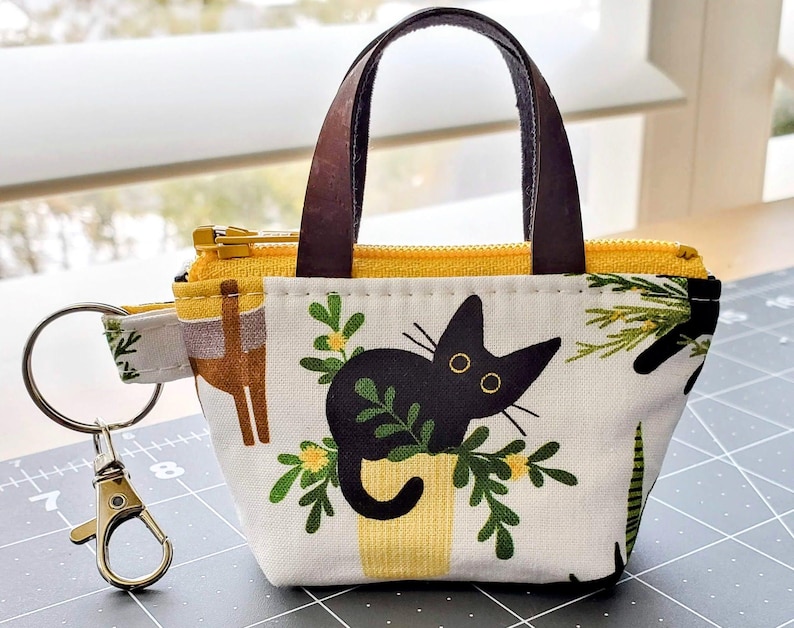 K&ouml;nnte beinhalten: Kleine Stoff-Tragetasche mit schwarzem Katzenmuster, gelbem Rei&szlig;verschluss und braunen Griffen. Die Tasche hat einen goldfarbenen Schl&uuml;sselanh&auml;nger und einen Stoffriemen mit Muster. Die Katze sitzt in einem gelben Topf mit gr&uuml;nen Bl&auml;ttern.