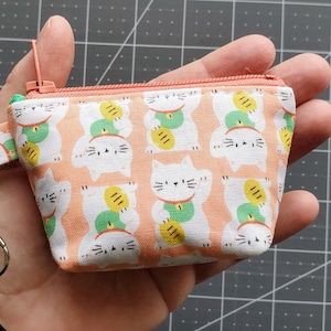 K&ouml;nnte beinhalten: Ein kleiner, rechteckiger M&uuml;nzbeutel mit pfirsichfarbenem Hintergrund und einem sich wiederholenden Muster aus wei&szlig;en Gl&uuml;ckskatzen, die Goldm&uuml;nzen halten. Der Beutel hat einen rosa Rei&szlig;verschluss und einen silbernen Schl&uuml;sselanh&auml;nger.