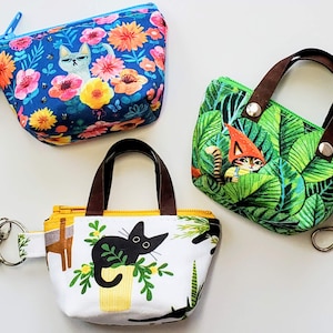 K&ouml;nnte beinhalten: Drei kleine, gemusterte Taschen mit Schl&uuml;sselanh&auml;ngern. Eine blaue Tasche hat ein Blumen- und Katzenmuster. Eine gr&uuml;ne Tasche hat ein Blatt- und Katzengnom-Design. Die dritte Tasche ist wei&szlig; mit einem schwarzen Katzen- und Pflanzenmuster.