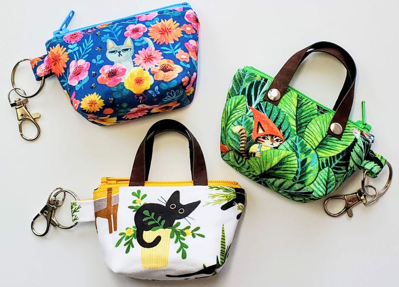 K&ouml;nnte beinhalten: Drei kleine, gemusterte Taschen mit Schl&uuml;sselanh&auml;ngern. Eine blaue Tasche hat ein Blumen- und Katzenmuster. Eine gr&uuml;ne Tasche hat ein Blatt- und Katzengnom-Design. Die dritte Tasche ist wei&szlig; mit einem schwarzen Katzen- und Pflanzenmuster.