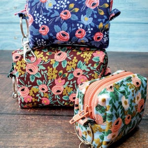 Puede incluir: Tres peque&ntilde;as bolsas de tela cuadradas con estampados florales. Las bolsas est&aacute;n apiladas, cada una con un dise&ntilde;o floral diferente en tonos azules, marrones y verdes, con flores rosas y melocot&oacute;n. Cada bolsa tiene una cremallera y un cierre met&aacute;lico.