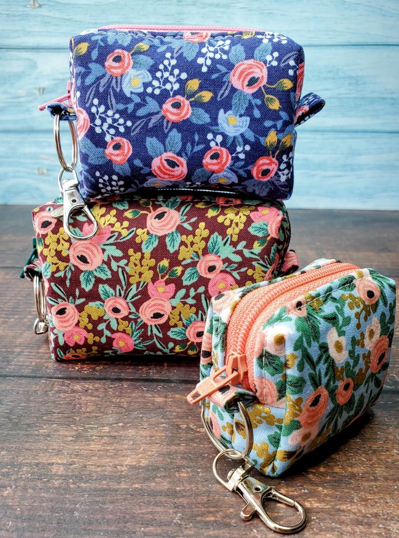 Puede incluir: Tres peque&ntilde;as bolsas de tela cuadradas con estampados florales. Las bolsas est&aacute;n apiladas, cada una con un dise&ntilde;o floral diferente en tonos azules, marrones y verdes, con flores rosas y melocot&oacute;n. Cada bolsa tiene una cremallera y un cierre met&aacute;lico.