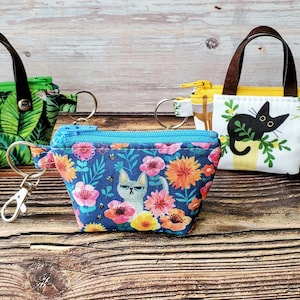 K&ouml;nnte beinhalten: Drei kleine, gemusterte M&uuml;nzb&ouml;rsen mit braunen Griffen und Schl&uuml;sselanh&auml;ngern. Eine ist gr&uuml;n mit einem Dschungelblattmuster, eine andere ist blau mit einem Blumen- und Katzenmuster, und die dritte ist wei&szlig; mit einer schwarzen Katzenillustration.