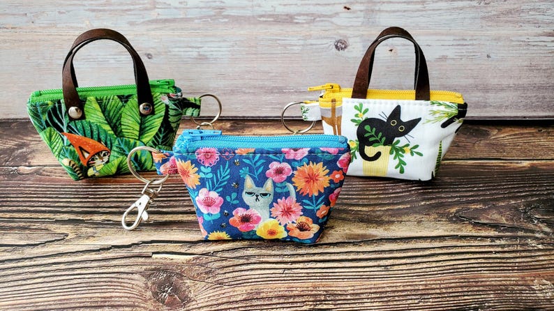 K&ouml;nnte beinhalten: Drei kleine, gemusterte M&uuml;nzb&ouml;rsen mit braunen Griffen und Schl&uuml;sselanh&auml;ngern. Eine ist gr&uuml;n mit einem Dschungelblattmuster, eine andere ist blau mit einem Blumen- und Katzenmuster, und die dritte ist wei&szlig; mit einer schwarzen Katzenillustration.