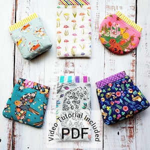 May include: An assortment of fabric pouches with various patterns, including foxes, mushrooms, flowers, birds, and rabbits. The pouches have colorful linings and are arranged on a white wooden surface. The text "Video Tutorial Included PDF" is visible.