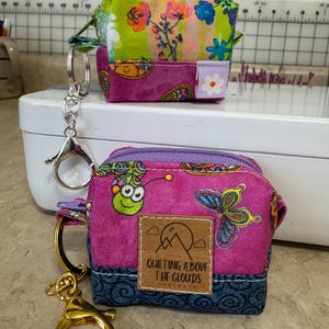 Puede incluir: Dos peque&ntilde;os monederos con cremallera y coloridos. Uno es verde y floral, el otro es rosa con una mariposa y un bicho verde. El monedero rosa tiene una etiqueta que dice "Quilting Above The Clouds". Ambos tienen llaveros.