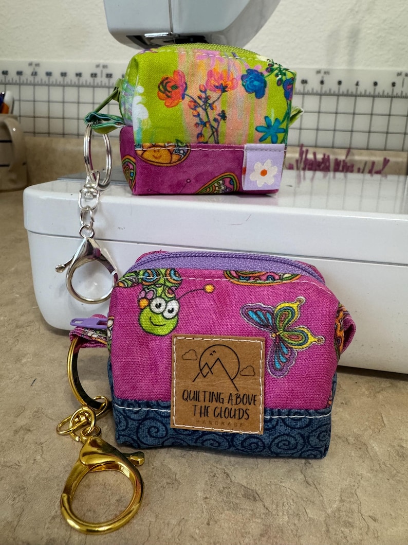 Puede incluir: Dos peque&ntilde;os monederos con cremallera y coloridos. Uno es verde y floral, el otro es rosa con una mariposa y un bicho verde. El monedero rosa tiene una etiqueta que dice "Quilting Above The Clouds". Ambos tienen llaveros.