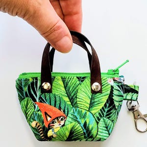 K&ouml;nnte beinhalten: Kleine, gr&uuml;ne Stoff-Tragetasche mit tropischem Bl&auml;ttermuster und braunen Ledergriffen. Die Tasche hat einen Rei&szlig;verschluss und einen Metallverschluss. Auf der Vorderseite ist eine Cartoon-Katze mit einem Zwergenhut aufgedruckt.