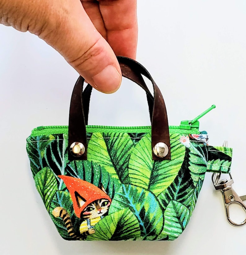 K&ouml;nnte beinhalten: Kleine, gr&uuml;ne Stoff-Tragetasche mit tropischem Bl&auml;ttermuster und braunen Ledergriffen. Die Tasche hat einen Rei&szlig;verschluss und einen Metallverschluss. Auf der Vorderseite ist eine Cartoon-Katze mit einem Zwergenhut aufgedruckt.