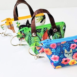 May include: Three small, patterned fabric purses with brown handles and metal key rings. The purses feature colorful designs including cats, flowers, and tropical foliage. Each purse has a different colored zipper.
