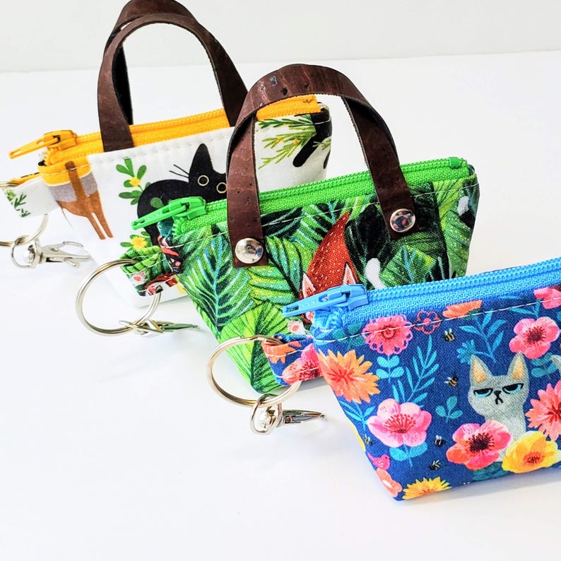 K&ouml;nnte beinhalten: Drei kleine Stoffb&ouml;rsen mit braunen Griffen und Metall-Schl&uuml;sselanh&auml;ngern. Die Geldb&ouml;rsen weisen farbenfrohe Muster auf, darunter Katzen, Blumen und tropisches Laub. Jede Geldb&ouml;rse hat einen Rei&szlig;verschluss in einer anderen Farbe.