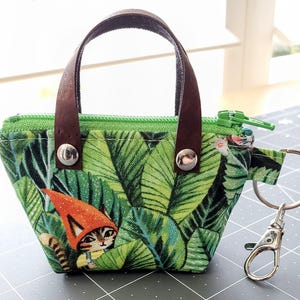 K&ouml;nnte beinhalten: Kleine Tragetasche aus gr&uuml;n gemustertem Stoff mit braunem Ledergriff. Die Tasche zeigt eine Katze mit einem Zwergenhut vor einem Hintergrund aus gr&uuml;nen Bl&auml;ttern. Ein silberner Verschluss und ein Schl&uuml;sselring sind seitlich angebracht.